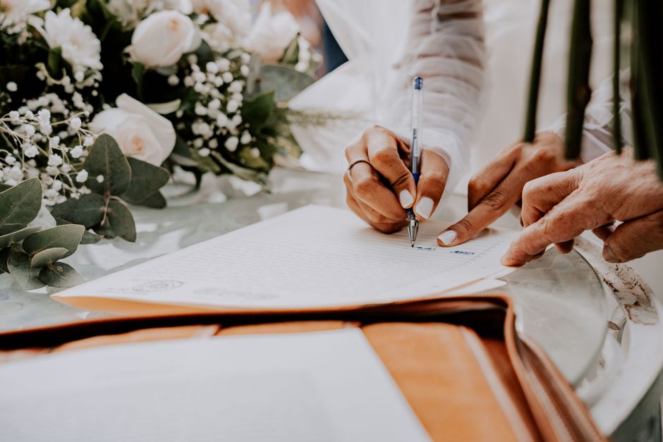 Close-up of a person signing a document with a blue pen, positioned on a table decorated with a bouquet of white roses, baby's breath, and eucalyptus leaves. The bouquet is arranged with delicate whit
