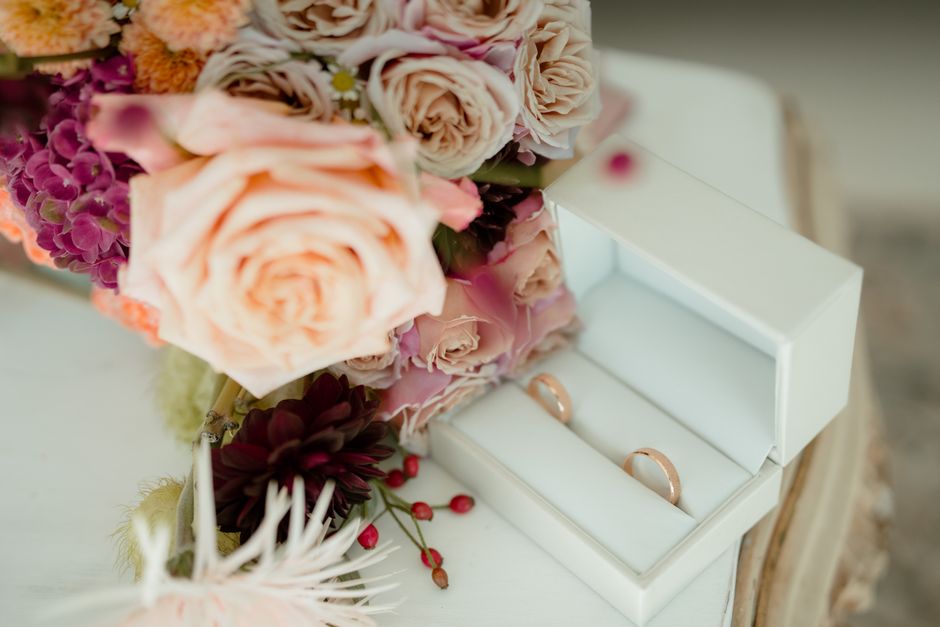 A close-up view of a bouquet featuring soft peach roses, pink and purple hydrangeas, and dark burgundy dahlias, arranged with fresh green foliage. The flowers are placed on a white surface, partially 