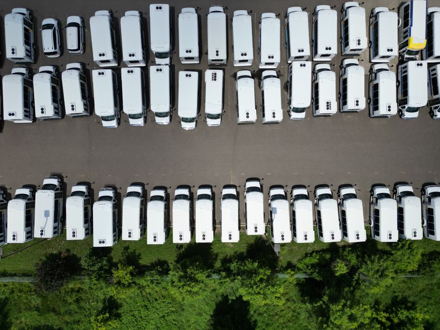 An aerial view of a parking lot filled with white delivery vans arranged in neat rows on a paved surface, with a strip of greenery and trees at the bottom of the image. The vans are parked side by sid