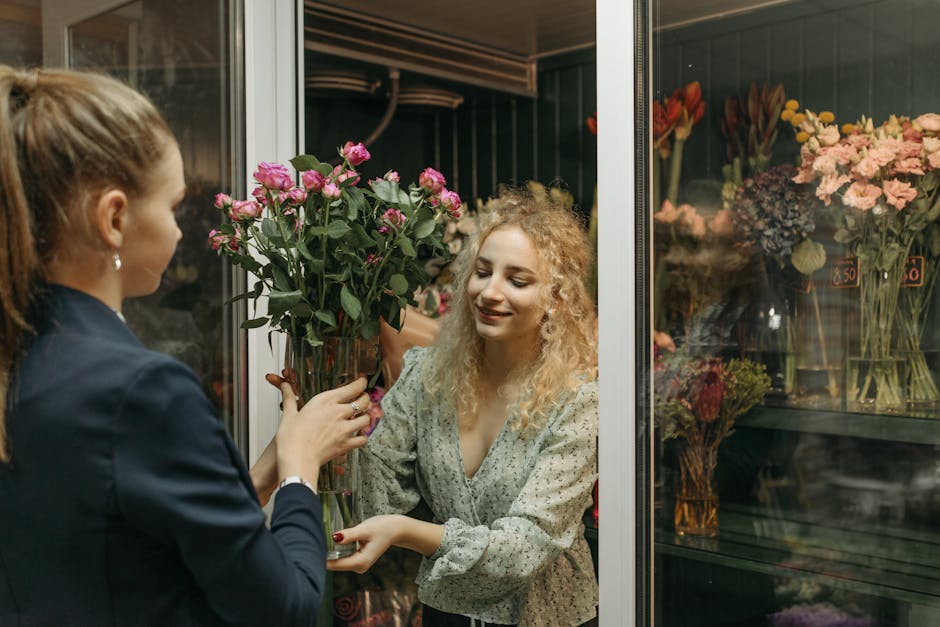 A florist with light brown hair tied back, dressed in a dark blazer, is handing a tall, clear glass vase filled with vibrant pink roses and lush green leaves to a smiling woman with curly blonde hair 
