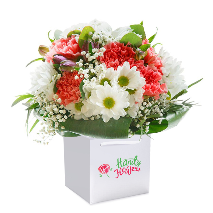 A tribute to Collier Row's love of fresh, uplifting florals, this bouquet is artfully arranged in a clean white square gift bag, its composition echoing the vibrancy of the surrounding neighbourhood. At the centre, coral-pink and red carnations unfurl their lavish ruffled petals in celebration, their lush hues complemented by clusters of white daisy-like chrysanthemums whose lime-green centers beam with lively charm-much like the friendly faces along Collier Row Road. Wisps of white baby's breath add a delicate, whimsical texture, softening the overall effect and lending a breath of innocence. Discreetly tucked among the blossoms, closed buds tinged with purple hint at new stories, evoking early morning walks past sunlit doorways. The bouquet is cradled by a ring of deep green, glossy foliage, providing both structure and a sense of natural abundance reminiscent of the green spaces sprinkled through the RM5 area. The ensemble forms an eye-catching, domed shape, every flower meticulously placed to radiate freshness and abundance. The sturdy bag, complete with elegant ribbon handles, is designed for easy gifting-ready to bring joy to a loved one's doorstep. Perfect for marking birthdays, anniversaries, or any occasion that deserves a special local gesture, this arrangement stands as a testament to Collier Row's passion for premium floristry and heartfelt connections.