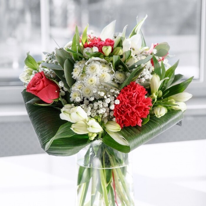 A refined red and white bouquet sits at the centre of the scene, arranged in a clear cylindrical vase that shows off every straight, freshly cut green stem beneath the waterline, emphasizing its just-prepared freshness. Above the glass, the flowers gather into a lush dome of colour and texture. At the heart are classic deep red roses, their velvety petals unfolding in layered spirals, partnered with a bold crimson carnation whose densely ruffled petals create a textured focal point at the front. These passionate reds are softened and balanced by an array of white blooms: graceful white Alstroemeria with delicate speckling on their inner petals, round white chrysanthemum or gerbera-style flowers, and several elegant white lily buds, still closed and pointing upwards, suggesting beauty yet to be revealed. Fine clouds of baby's breath weave between the larger blossoms, giving the arrangement a light, airy finish and a sense of celebration. The base is thoughtfully framed by broad, glossy dark green leaves wrapped and curved into place, almost like a sculpted collar, while additional foliage in multiple green hues-slender pointed leaves and eucalyptus-like stems-adds layered depth. The bouquet is photographed on a spotless white surface, with a softly lit, out-of-focus background that could be a bright front room or hallway in Collier Row, filled with natural daylight. The overall mood is polished yet warm, ideal as a centrepiece for a family gathering off Collier Row Lane, a romantic gesture delivered locally, or a thoughtful gift that brings both elegance and comfort into any home or office setting.