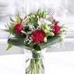A luxurious red and white bouquet stands proudly in a clear cylindrical glass vase, its freshly cut green stems visible through the water and neatly aligned, giving a sense of care and craftsmanship. Deep red roses, their velvety petals beginning to unfurl, sit alongside a richly ruffled crimson carnation at the front, creating a passionate focal point that immediately captures the eye. Around them, elegant white Alstroemeria with fine speckles, creamy white lily buds still tightly closed, and soft white chrysanthemums and gerbera-style blooms bring a feeling of purity and calm. Wisps of delicate baby's breath float between the larger flowers like tiny stars, offering a cloud-like softness. The base of the bouquet is wrapped in broad, glossy dark green leaves, carefully folded and curved to form a sculpted collar that frames the blossoms. Additional foliage in varied shades of green, including eucalyptus-like sprigs and pointed leaves, adds movement, texture, and depth. The arrangement is photographed on a spotless white surface against a gently blurred, light-filled background that could be a bright kitchen or lounge in Collier Row, the natural daylight making every petal glow with freshness. The overall impression is of a polished, modern take on classic romance, ideal as a centrepiece for an anniversary dinner on Havering Road, a birthday surprise delivered to a Collier Row office, or simply to bring warmth and admiration into a family home, radiating affection and quiet celebration.