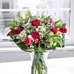 A lush, romantic bouquet of red roses, red carnations, white alstroemeria, and soft clouds of baby's breath sits proudly in a clear glass vase on a bright white table, bathed in gentle natural light from a large window. The crimson roses are the immediate focal point, some fully open with velvety, layered petals curling outward, others still in smooth, elegant buds that promise to unfurl over the next few days. Around them, ruffled red carnations add depth and texture, their densely packed petals creating a sense of fullness and abundance. Threaded between the richer reds are crisp white alstroemeria, many still in bud, their slender green-tipped petals just beginning to part, alongside a few partially open blooms that reveal delicate, trumpet-shaped flowers with soft markings deep within. Tiny white gypsophila, or baby's breath, is sprinkled throughout like a mist of little stars, softening the arrangement and lending a dreamy, airy feel. The flowers are cradled by an array of glossy green foliage: broad, deep green leaves wrap around the base in a structured collar, while slimmer, lighter stems weave through the centre, adding movement and contrast. Through the crystal-clear vase, the neatly trimmed stems are visible, immersed in fresh water that catches hints of reflected light. Behind the arrangement, the view through the window is muted and slightly blurred, showing a pale, almost monochrome outdoor scene that makes the bouquet's reds, whites, and greens appear even more vivid. It feels like a joyful, elegant display you might find on a dining table in a Collier Row home, perfect for a birthday, anniversary, or a simple, heartfelt thank-you delivered locally on a bright morning.